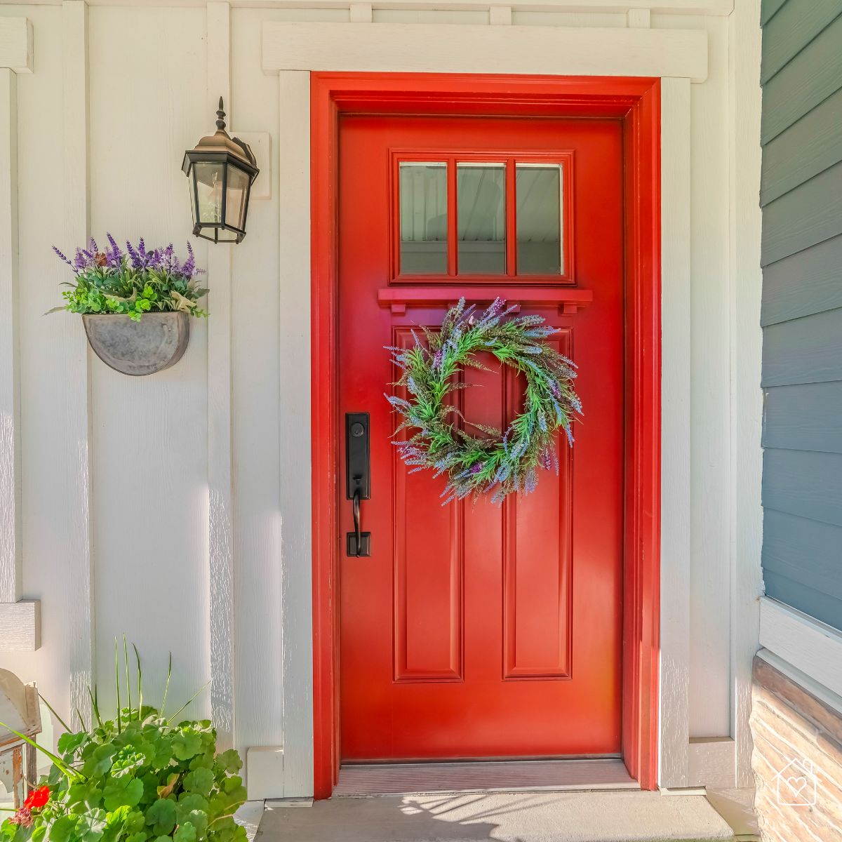 Vibrant red front door decorated with a green wreath, flanked by white siding and outdoor plants, exposed to the elements.