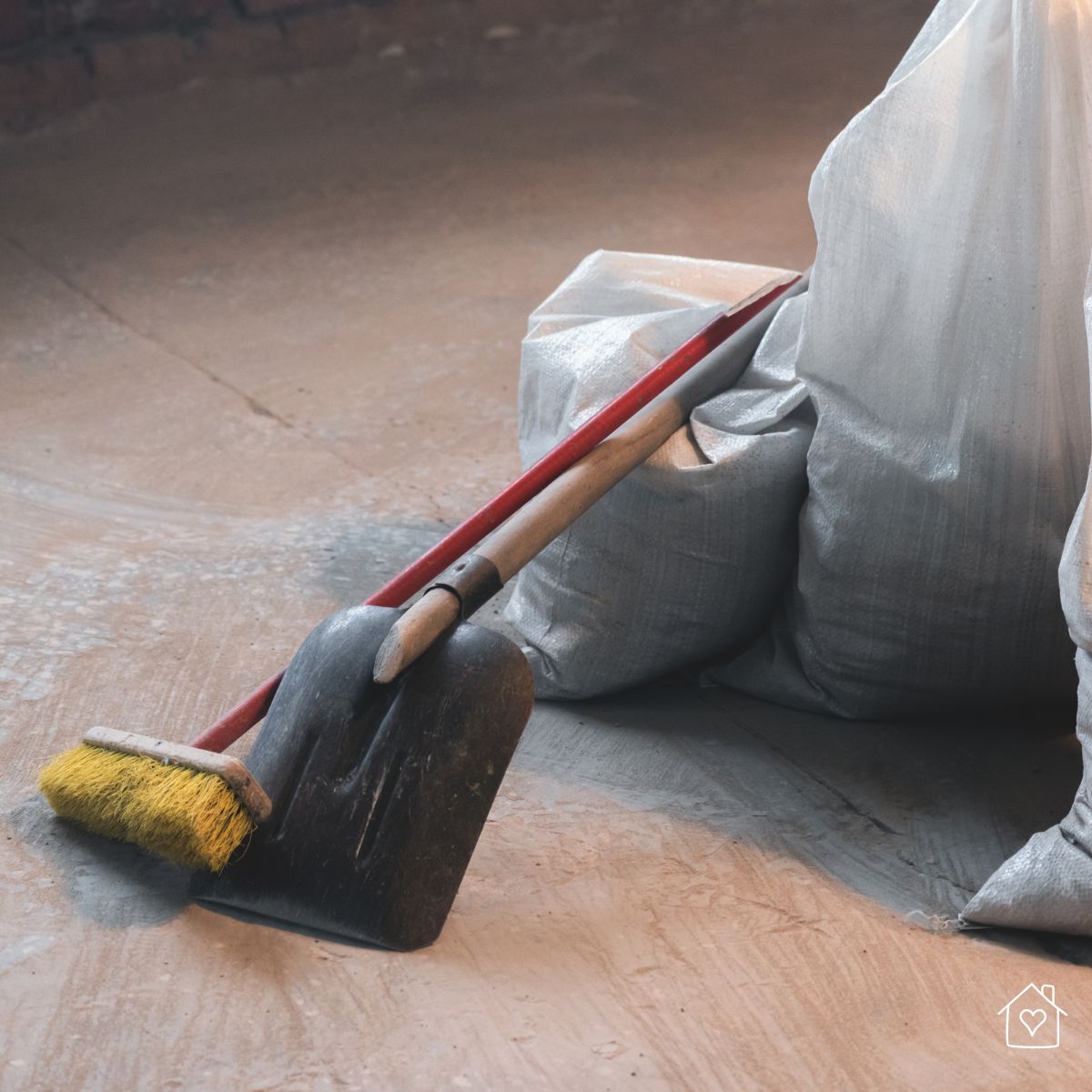 Close-up of a broom and shovel leaning against bags of construction debris on a dusty floor after renovation.