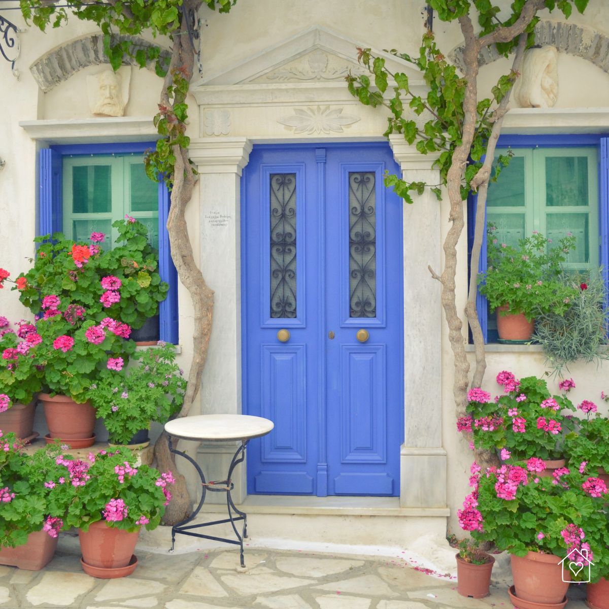 Bright blue double front doors with decorative ironwork, surrounded by potted pink flowers, showcasing a colorful yet weathered entrance.