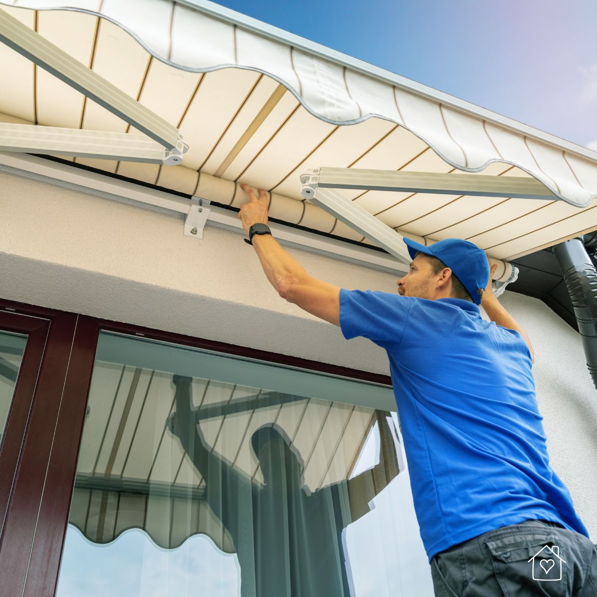 Installer in a blue uniform adjusting a white and tan striped retractable awning over a glass patio door.