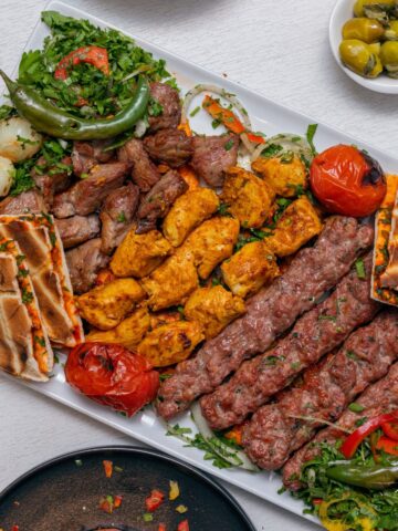 A large platter of grilled meats including chicken, lamb, and kofta kebabs, served with flatbread, grilled vegetables, and fresh herbs.