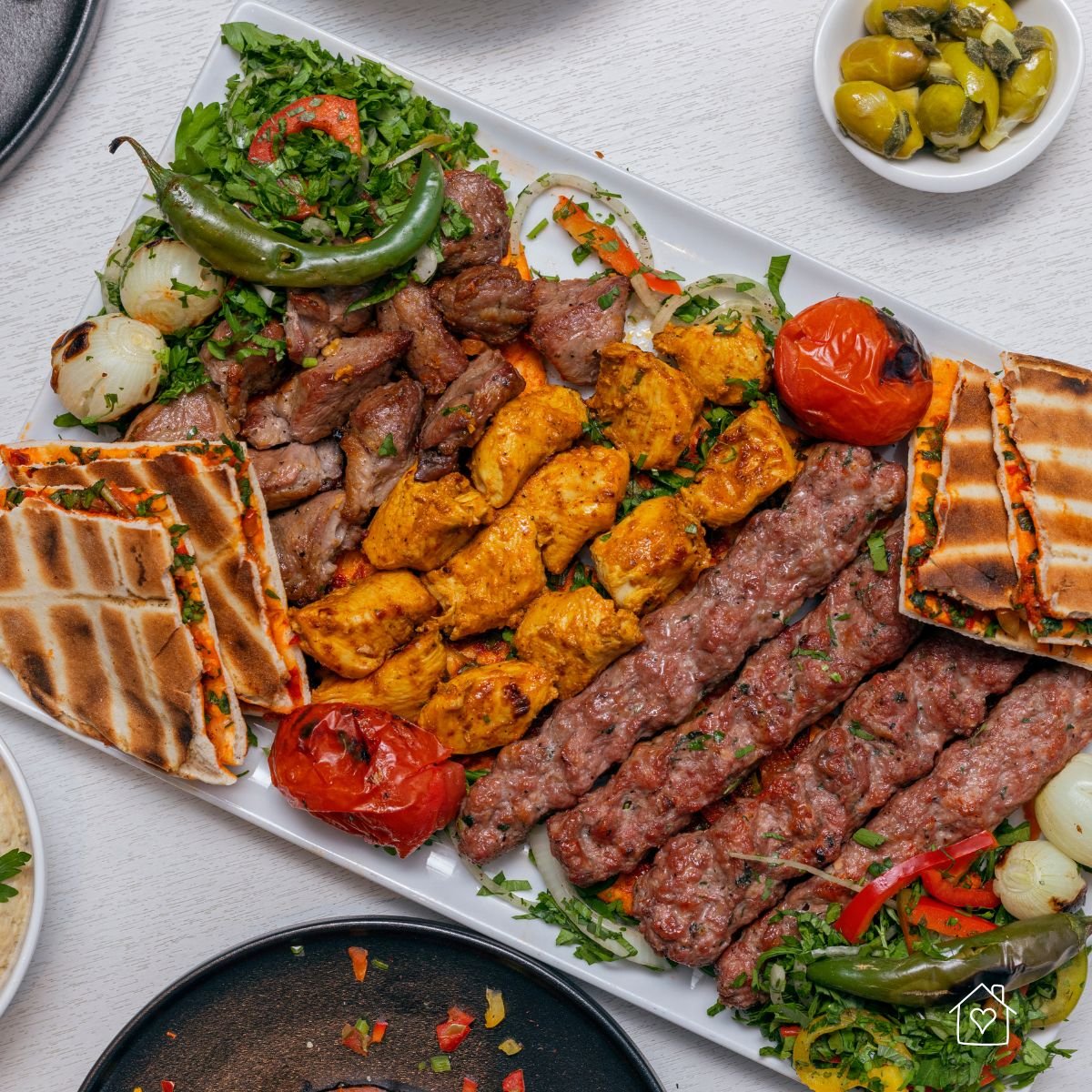 A large platter of grilled meats including chicken, lamb, and kofta kebabs, served with flatbread, grilled vegetables, and fresh herbs.