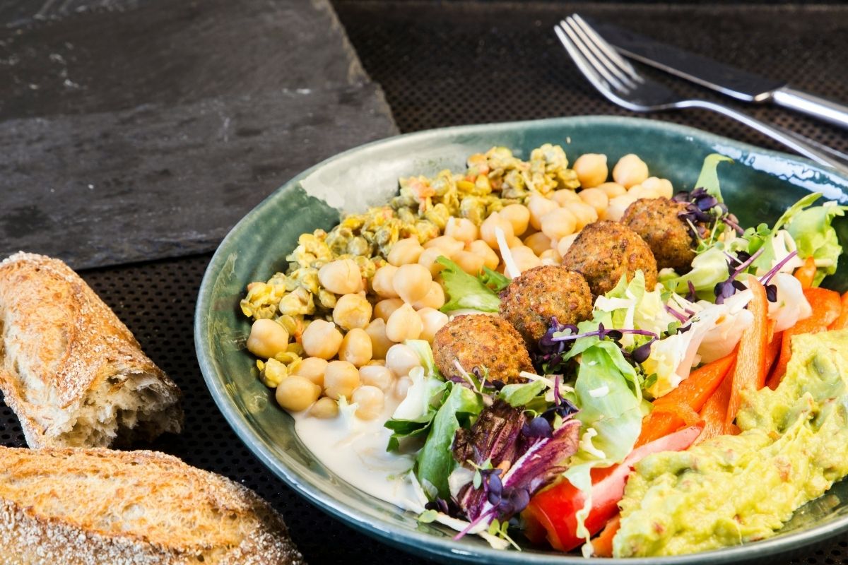 A hearty falafel bowl featuring crispy falafel, chickpeas, fresh greens, guacamole, and creamy yogurt dressing, served with crusty bread.
