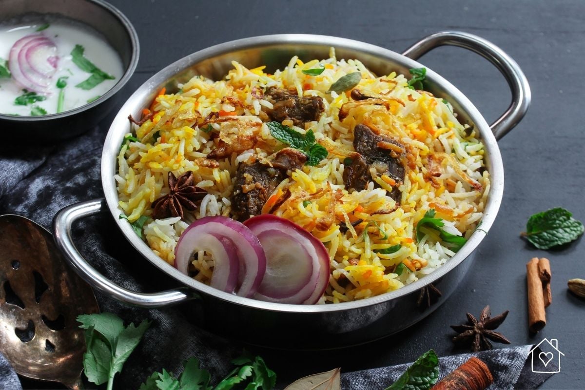A metal pot filled with colorful biryani rice, spiced meat, fried onions, and fresh herbs, accompanied by a side of yogurt and red onions.