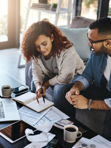 A young couple sitting together at a coffee table with a laptop, receipts, and paperwork while discussing personal loan options.