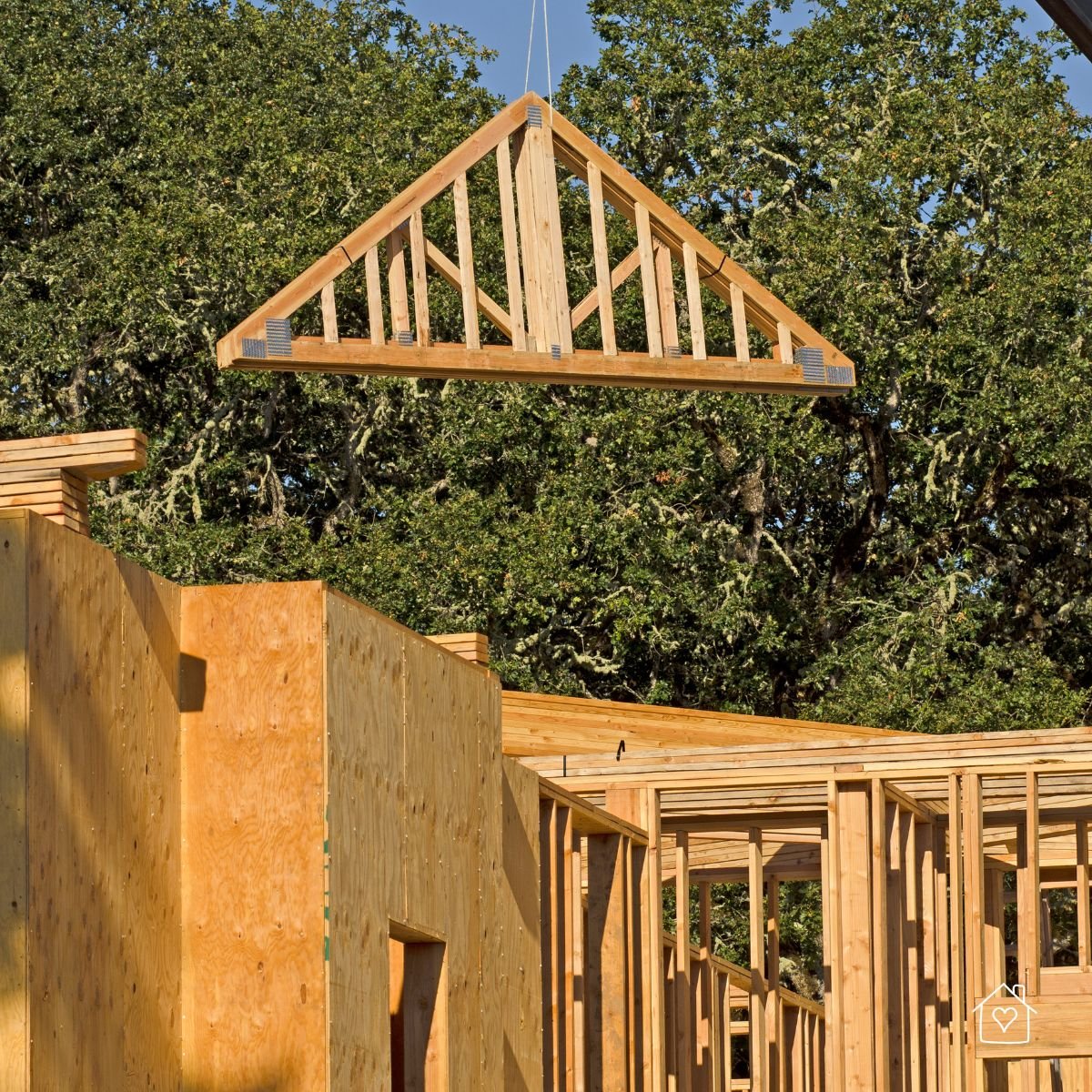 A wooden roof truss being positioned on top of framed walls during a DIY home construction project, surrounded by trees.