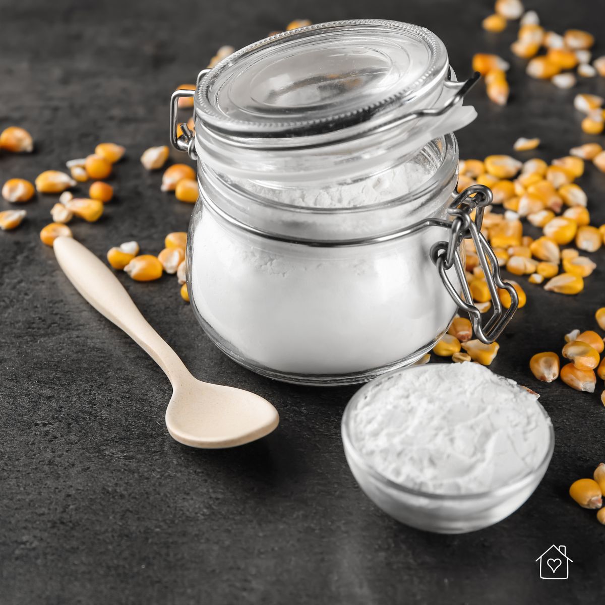 A jar and bowl of cornstarch with a spoon, surrounded by corn kernels&mdash;great for treating oily carpet spots naturally.