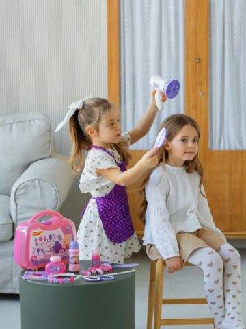 Two young girls pretending to run a hair salon at home, using a toy blow dryer and hairbrush.