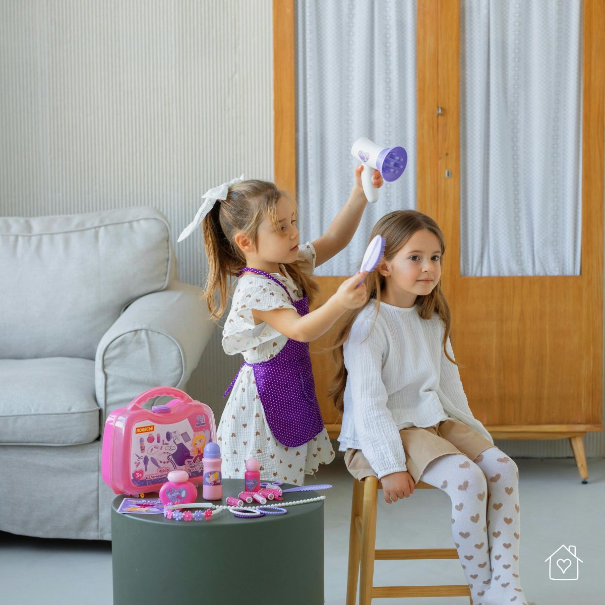 Two young girls pretending to run a hair salon at home, using a toy blow dryer and hairbrush.