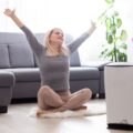 Smiling woman stretching with arms up while sitting near an air purifier in a cozy living room.