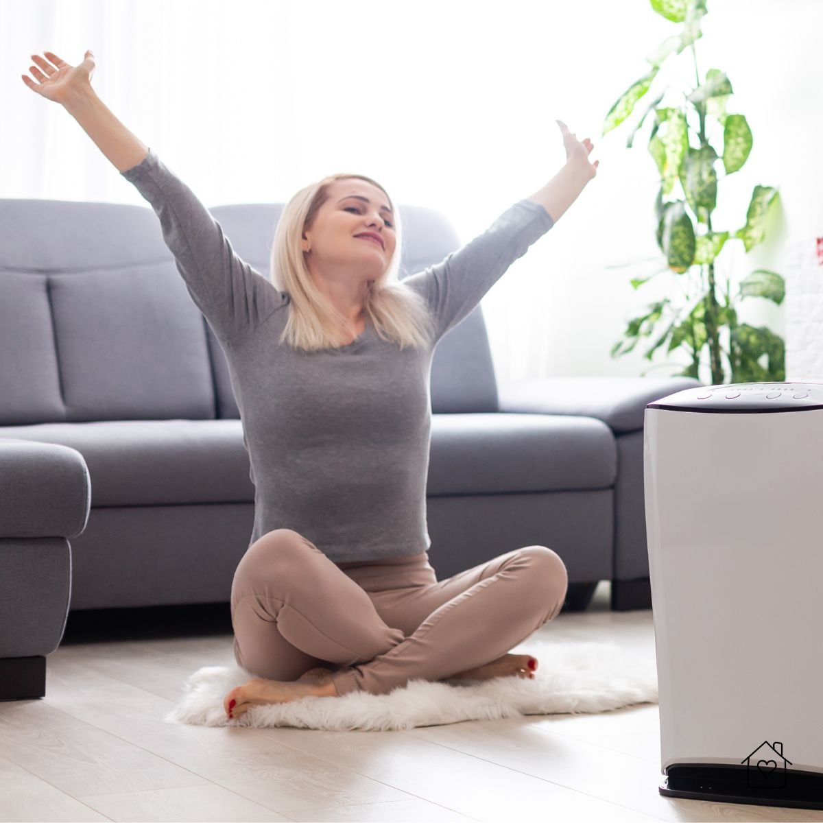 Smiling woman stretching with arms up while sitting near an air purifier in a cozy living room.