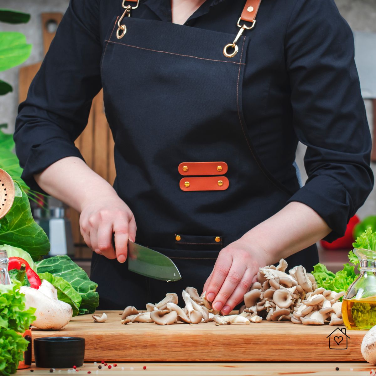 A home cook wearing a black apron chopping fresh mushrooms on a wooden cutting board with a Japanese santoku knife.