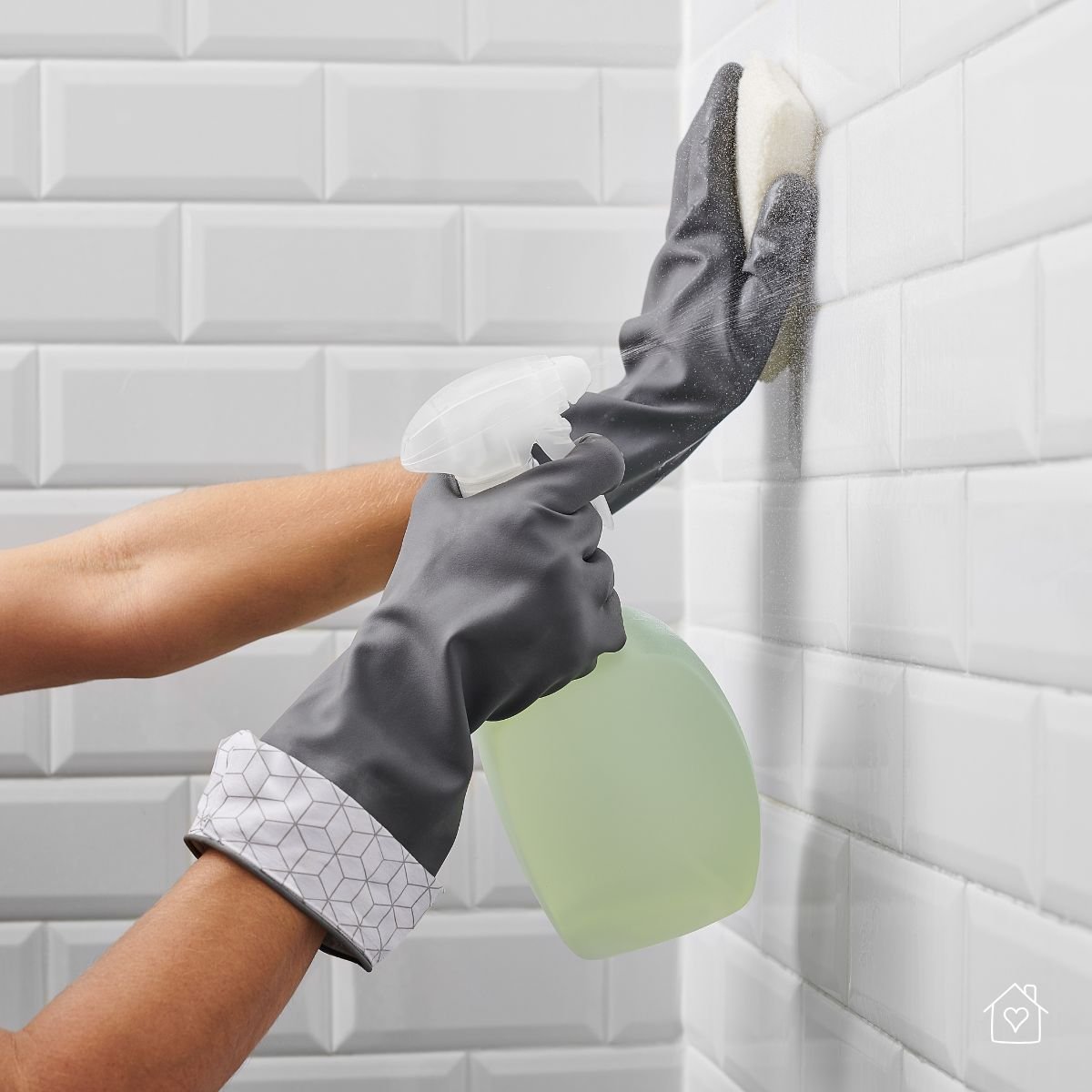 Hands wearing gray cleaning gloves scrubbing white bathroom tiles with a sponge and spray bottle.