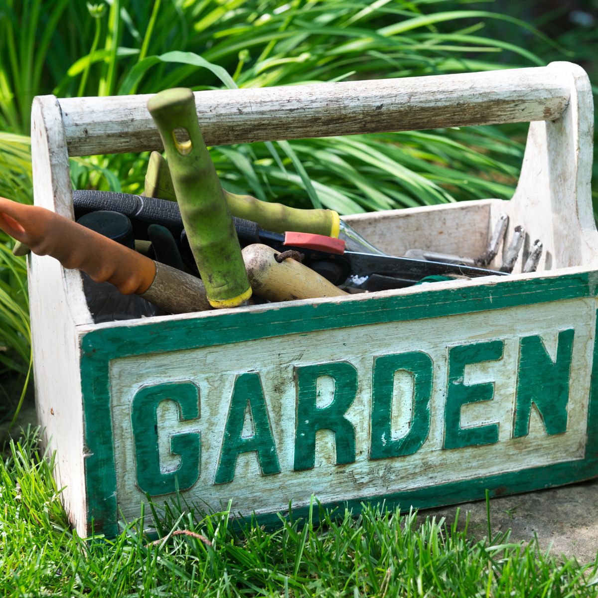 Rustic wooden garden crate holding hand tools with green handles, sitting on grass.