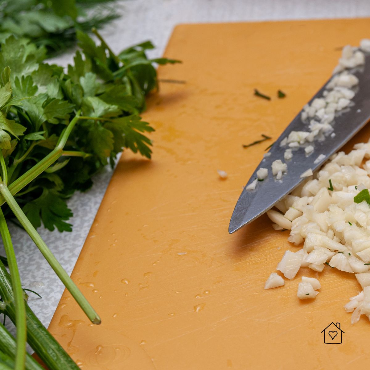 A sharp kitchen knife with minced garlic on a cutting board, fresh parsley beside it for cooking preparation.