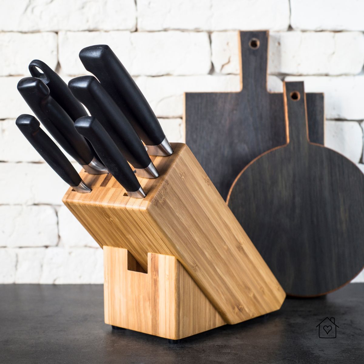 A set of professional kitchen knives in a wooden block with dark cutting boards against a white brick background.