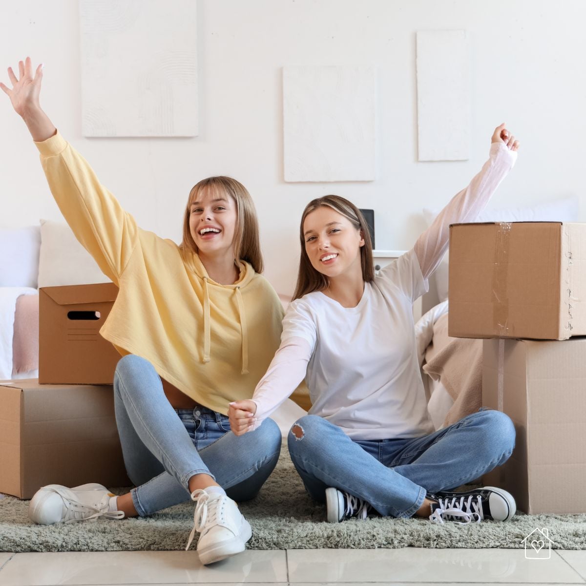 Two smiling young women sitting on the floor surrounded by moving boxes while decluttering together.