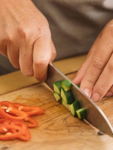 A close-up of hands slicing cucumber cubes and red pepper rings on a wooden cutting board with a sharp knife.
