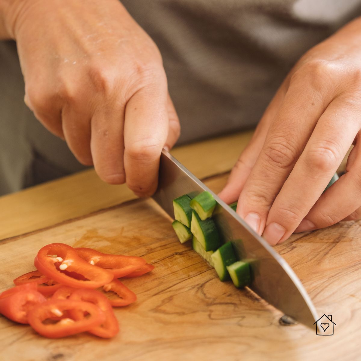 A close-up of hands slicing cucumber cubes and red pepper rings on a wooden cutting board with a sharp knife.