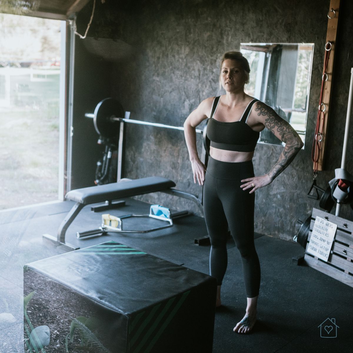 Woman standing confidently in a garage gym with a barbell bench and exercise equipment.