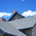 Gable roof with ridge cap, attic vent, and brick chimney against a bright blue sky.