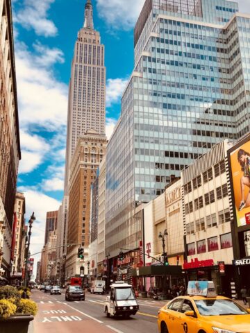 Street view of New York City with the Empire State Building and modern glass high-rise buildings, yellow taxi in the foreground, and busy shops along the avenue.