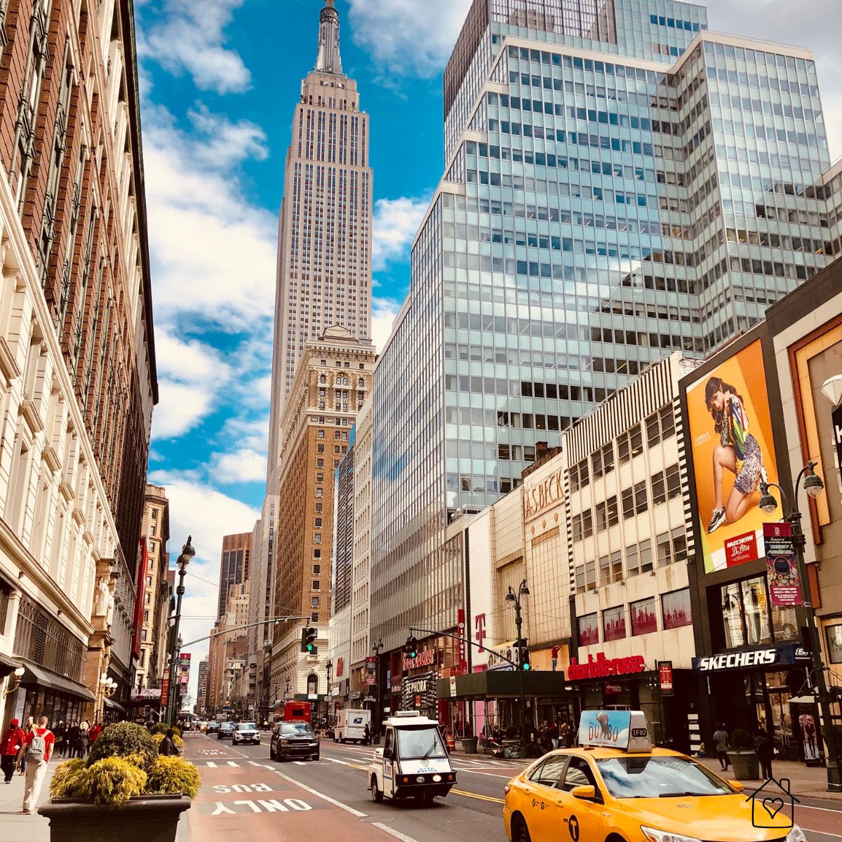 Street view of New York City with the Empire State Building and modern glass high-rise buildings, yellow taxi in the foreground, and busy shops along the avenue.