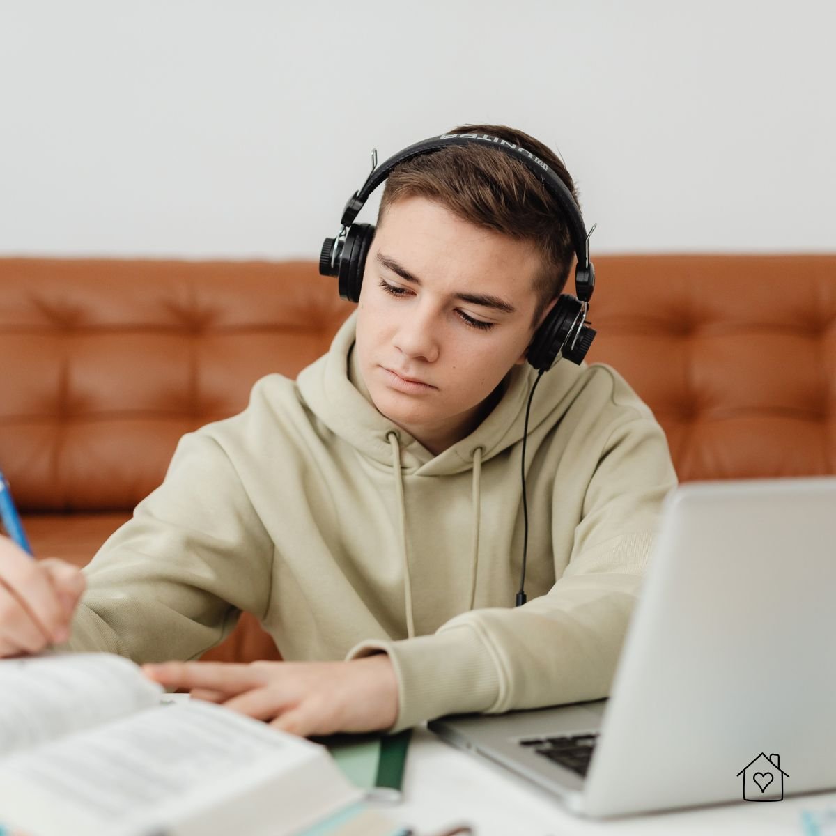 Teen boy wearing headphones studying at home with a laptop and notebook open, focused on writing during a homeschool lesson.