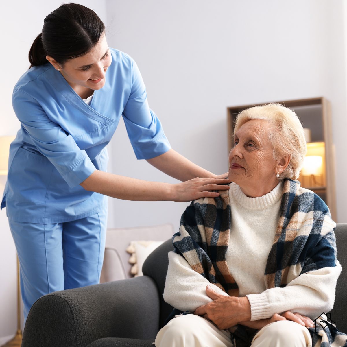 Caregiver in blue scrubs offering support to an older woman sitting on a couch, symbolizing compassionate senior care and guidance when choosing a nursing home.