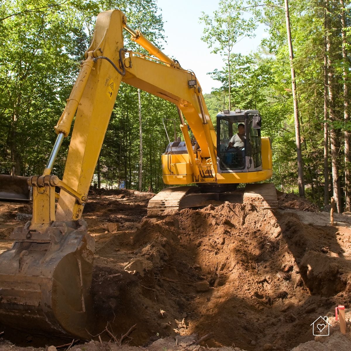 Yellow crawler excavator digging a deep trench on a wooded lot with fresh soil piles around the machine.