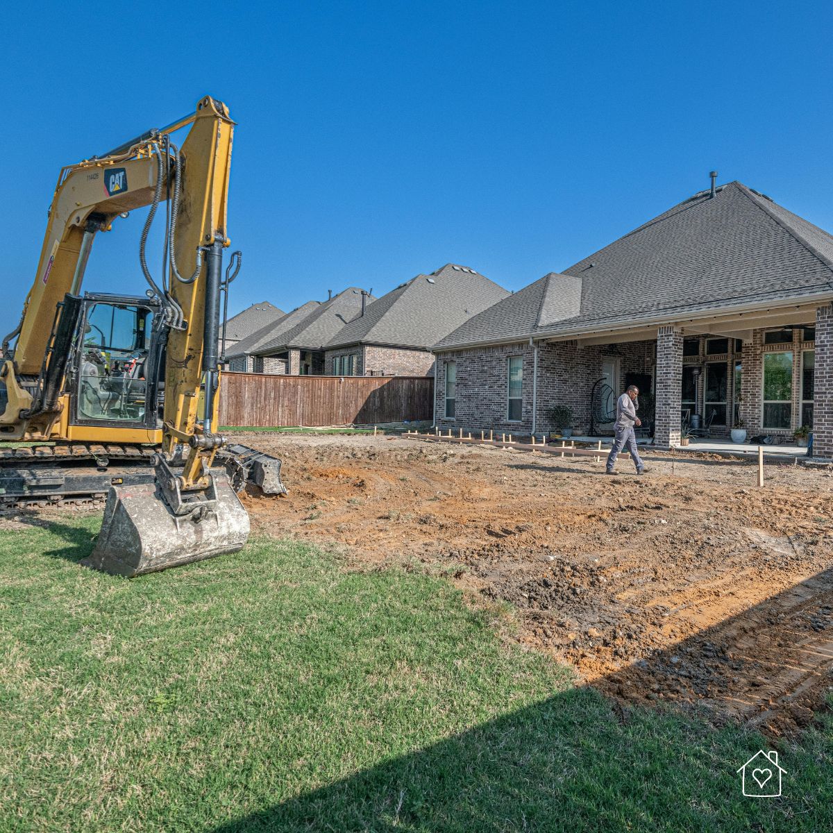 Compact excavator grading a suburban backyard behind brick homes, with forms set for a small foundation.