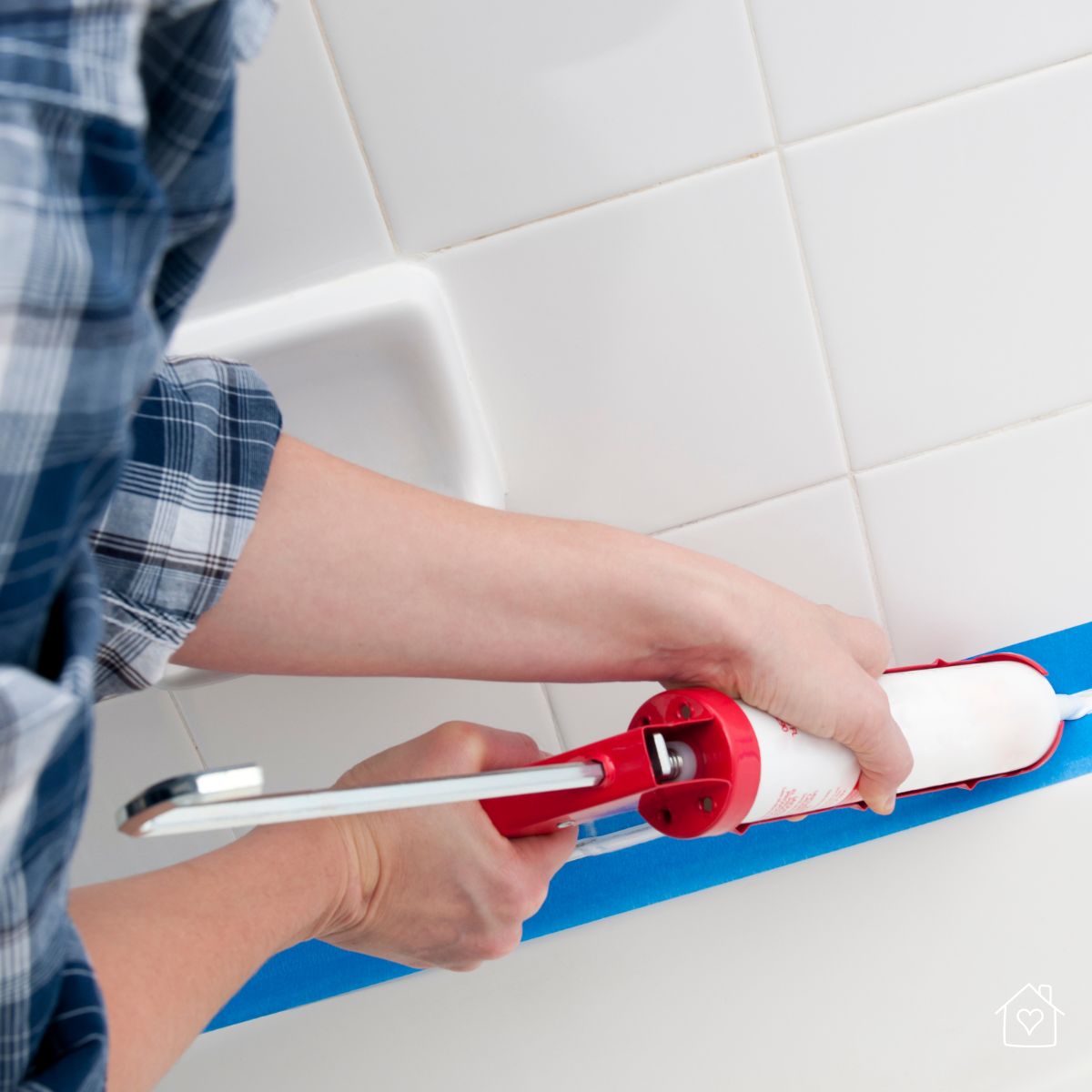 Hands apply fresh white caulk along the tub and tile edge using a caulk gun and painter’s tape.