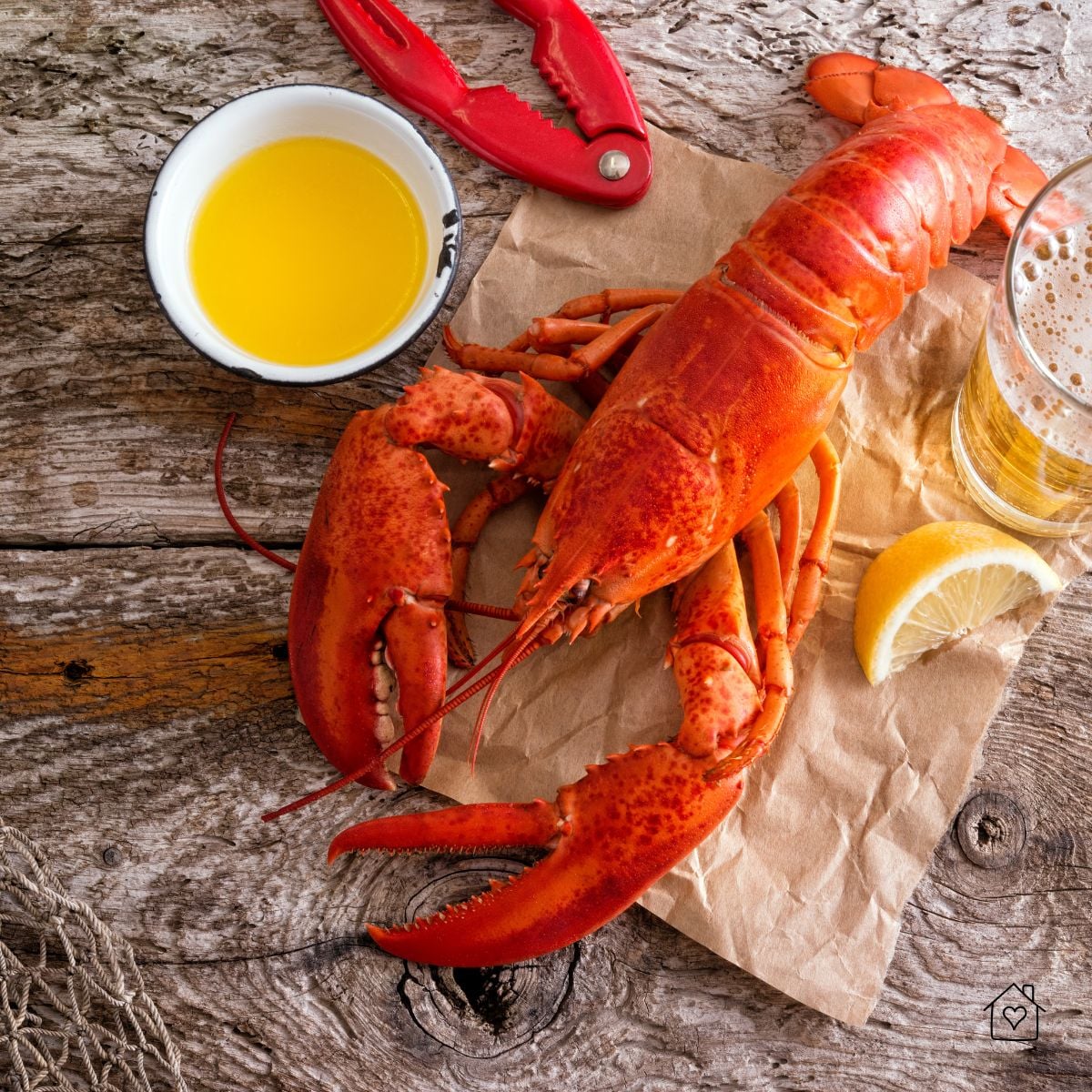 A bright red cooked lobster on brown paper with melted butter, lemon wedge, and beer on a rustic wooden table.