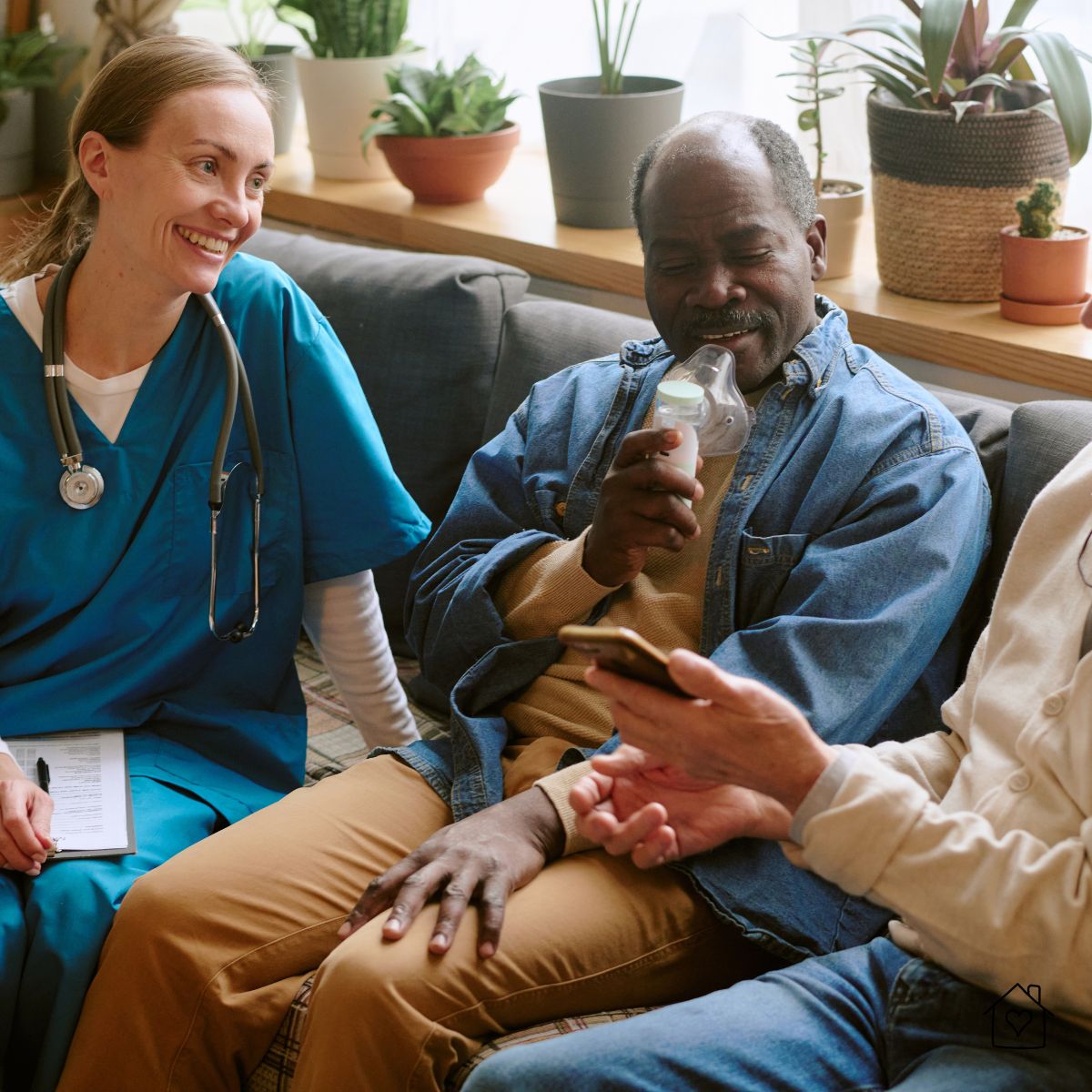 Smiling nurse supporting an older adult using a nebulizer at home while a loved one shows him something on a phone.