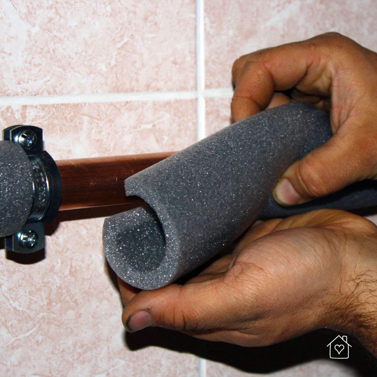 Hands sliding foam insulation over an exposed copper pipe on a tiled wall to prevent winter freezes and heat loss.