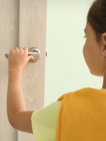 A young girl wearing a yellow shirt opens a light-colored interior door, emphasizing the need for supervision and childproof locks inside the home.