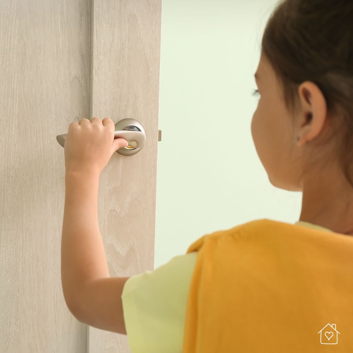 A young girl wearing a yellow shirt opens a light-colored interior door, emphasizing the need for supervision and childproof locks inside the home.