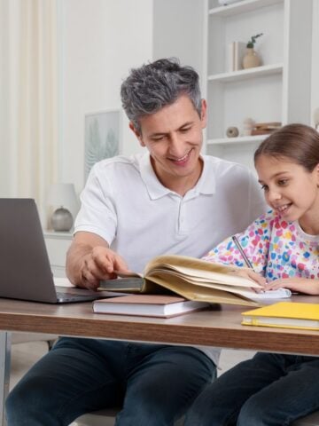 Father and daughter sitting together at a desk during a homeschool lesson, smiling as they review a writing assignment with books and a laptop nearby.
