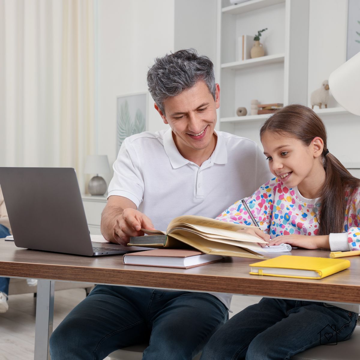 Father and daughter sitting together at a desk during a homeschool lesson, smiling as they review a writing assignment with books and a laptop nearby.