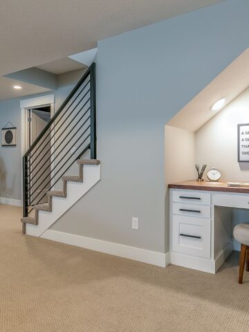Small basement workspace tucked beneath a staircase, featuring a white built-in desk, soft lighting, and a motivational wall quote for a functional home office nook.