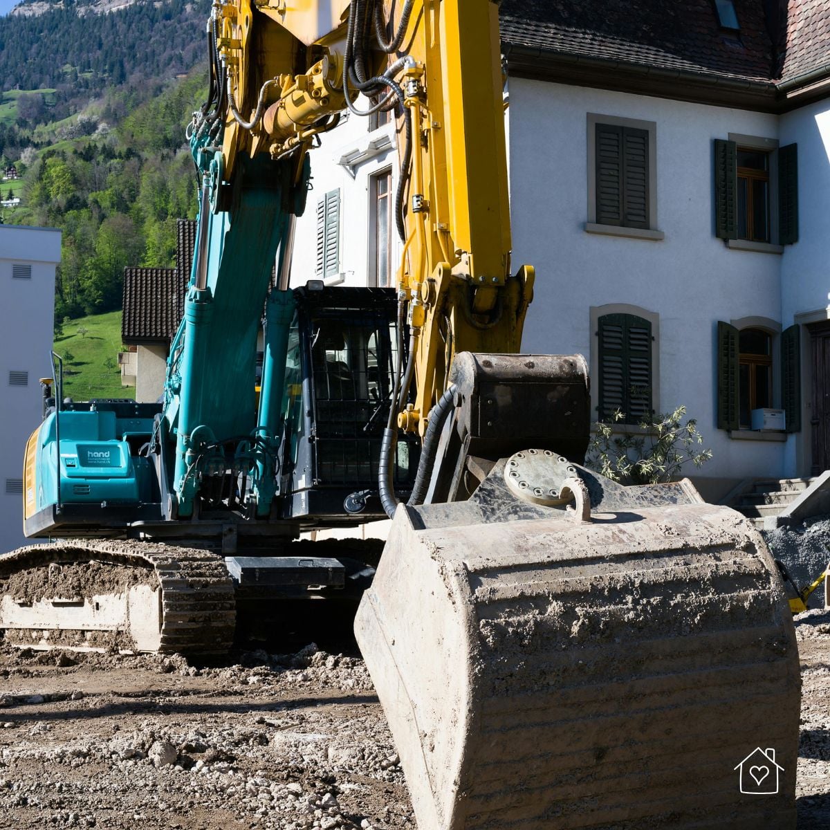 Excavator operating close to a house along a narrow street, showing space constraints near existing structures.