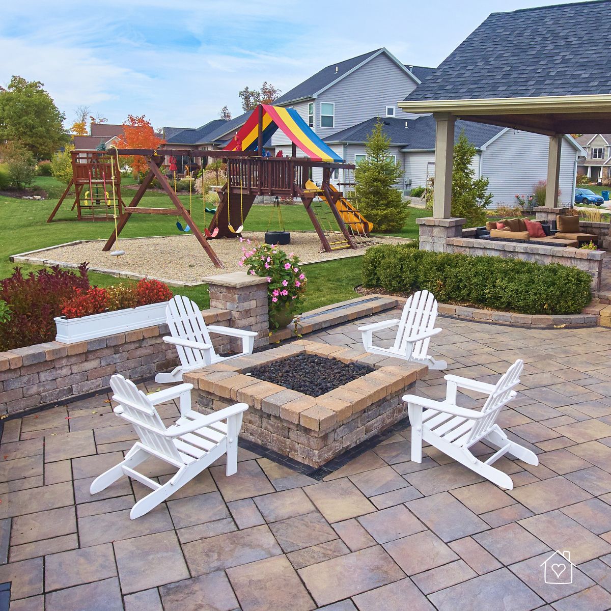 Square stone fire pit on a paver patio with white Adirondack chairs; playset and covered seating area in the background.