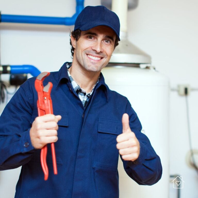 Smiling plumber in a navy uniform holding a pipe wrench and giving a thumbs-up beside a home water heater-professional help nearby.