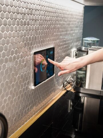 Hand tapping a wall-mounted tablet controller on a hex-tile kitchen backsplash, showing a thermostat/energy app beside a coffee machine and cooktop.