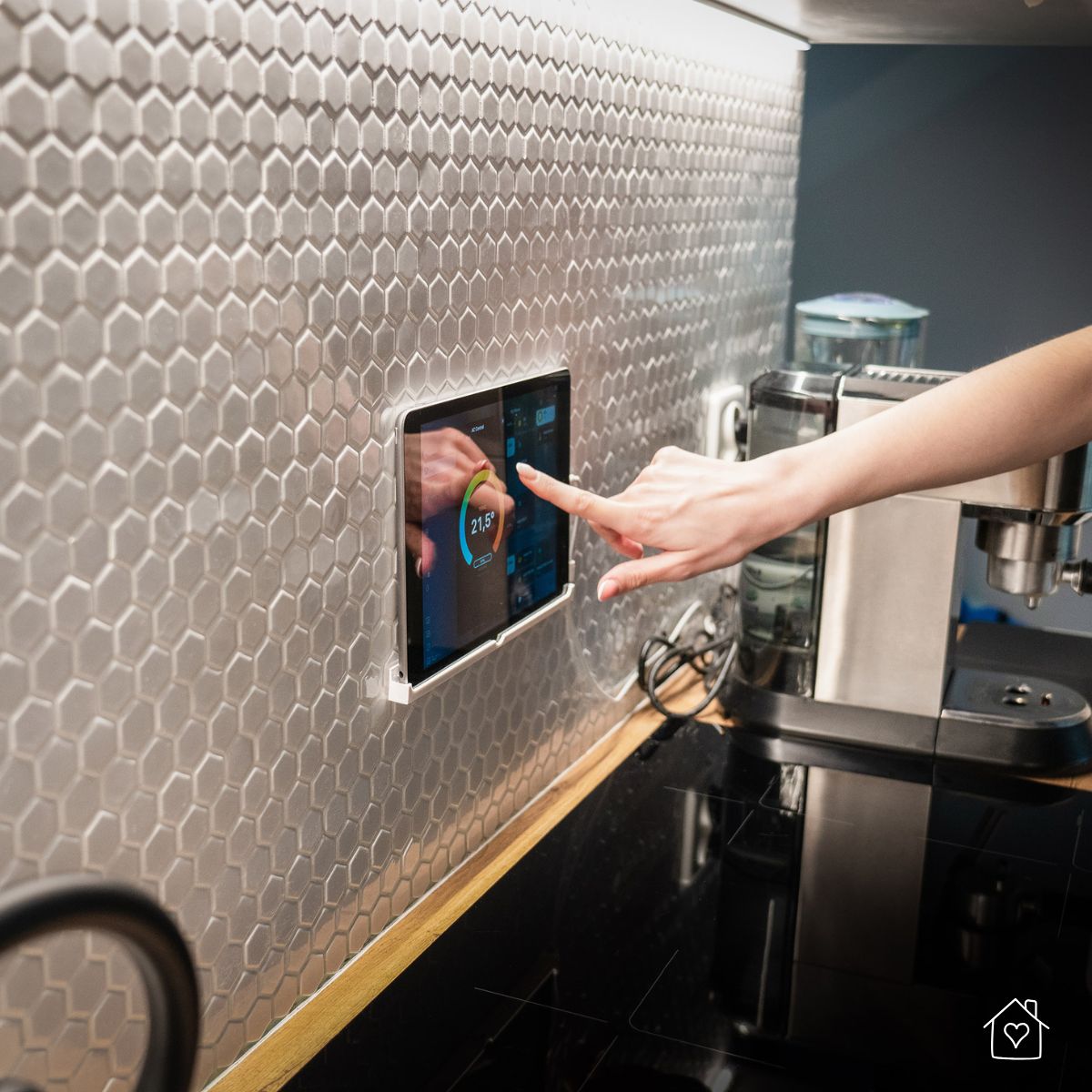 Hand tapping a wall-mounted tablet controller on a hex-tile kitchen backsplash, showing a thermostat/energy app beside a coffee machine and cooktop.