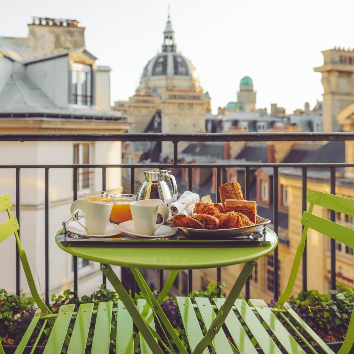 Green bistro table set with coffee, pastries, and juice on a Paris balcony, classic rooftops and a domed building in soft morning light.