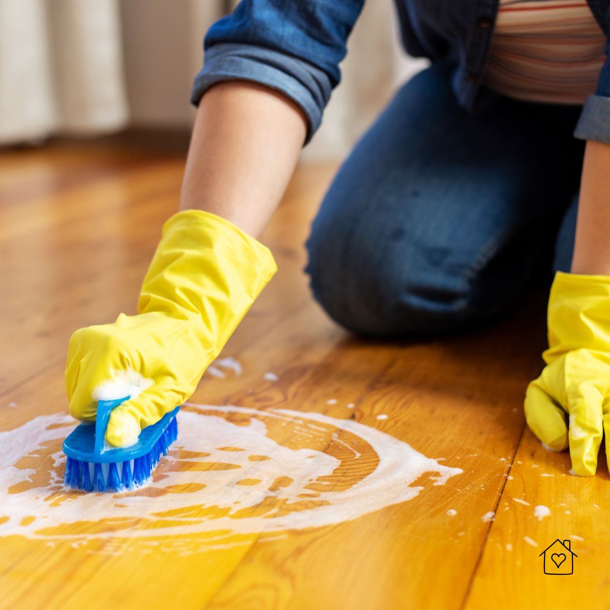 Hands in yellow rubber gloves scrubbing a hardwood floor with a blue brush and soapy water.