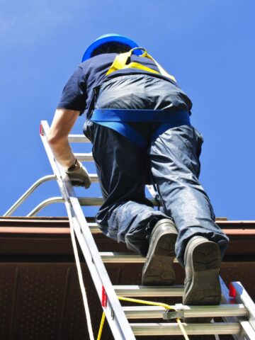 Person climbing an extension ladder to a roof while wearing a helmet and harness, demonstrating safe roof access.
