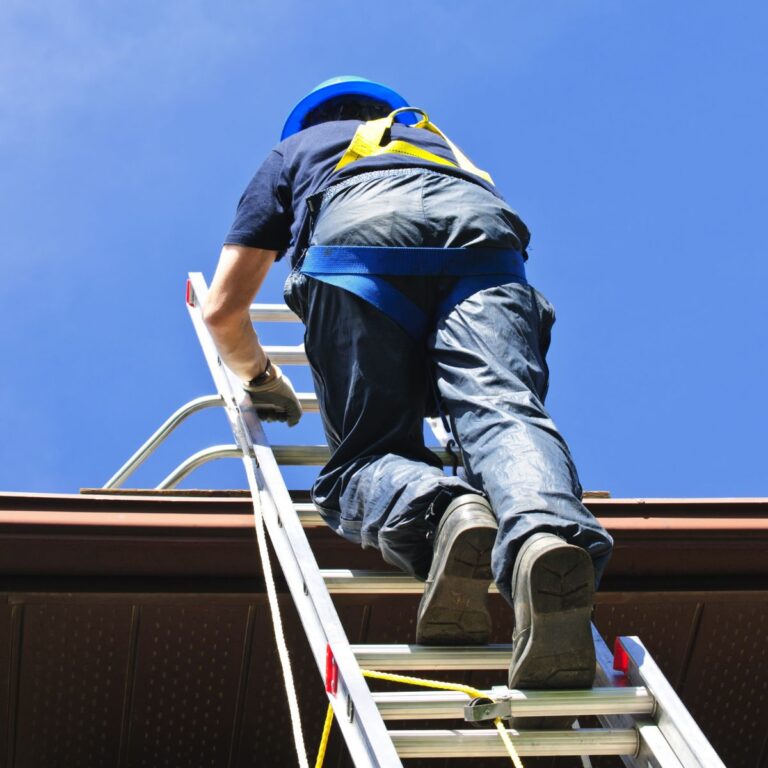 Person climbing an extension ladder to a roof while wearing a helmet and harness, demonstrating safe roof access.
