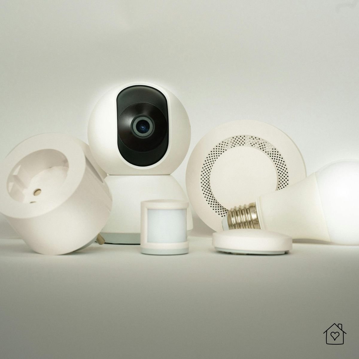 Group of white smart home gadgets including a security camera, motion sensor, smart plug, and smart light bulb arranged on a neutral surface.